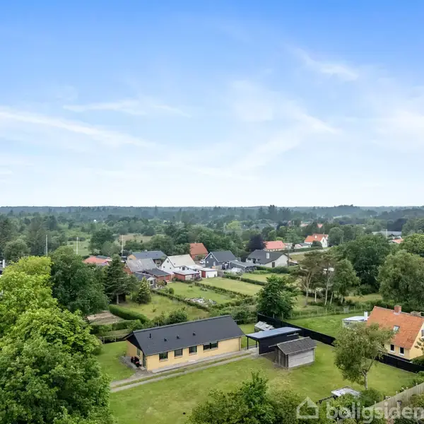 Landsbyhuse ligger spredt i et grønt, frodigt område med træer og marker. En blå himmel med lette skyer danner baggrund ovenover.