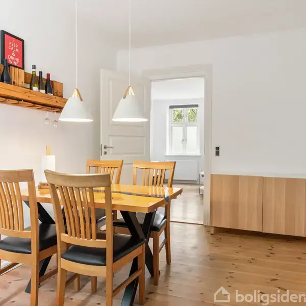 En spisestue med et træbord omgivet af fire stole. Bordet er placeret under to hængelamper. På væggen er en vinreol med flasker og et skilt med teksten "Keep calm and drink wine". I baggrunden ses en åbning til et andet rum med vinduer.