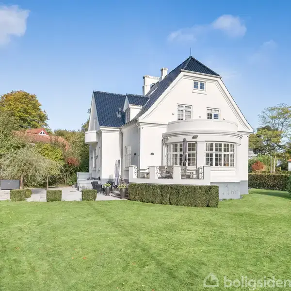 Et stort hvidt hus med gråt tag står i en velplejet have med klippede buske og åbent græsareal. Huset har en terrasse med havemøbler.