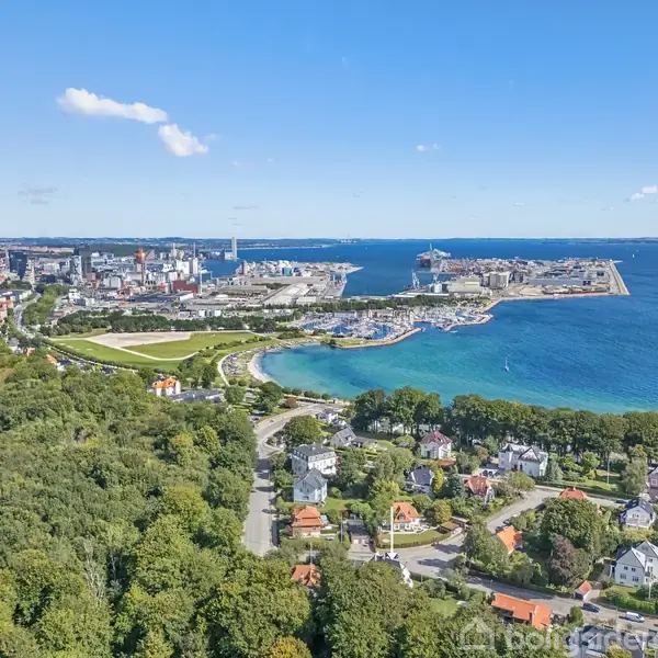 Byområde ved kysten ses oppefra, hvor boliger omgives af træer i forgrunden. En havn strækker sig mod horisonten, og et klart blåt hav omkranser landskabet under en blå himmel.