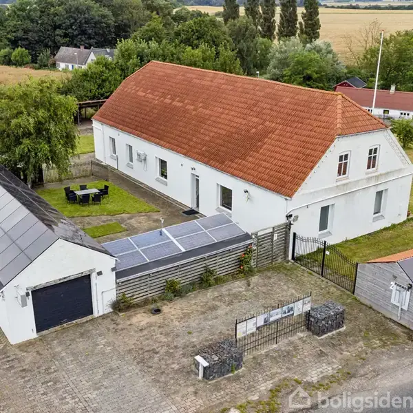 Et hvidt landhus med rødt tag omkranset af en flisebelagt gårdsplads. Solpaneler er installeret på taget af en tilhørende bygning. Omgivet af marker og træer.