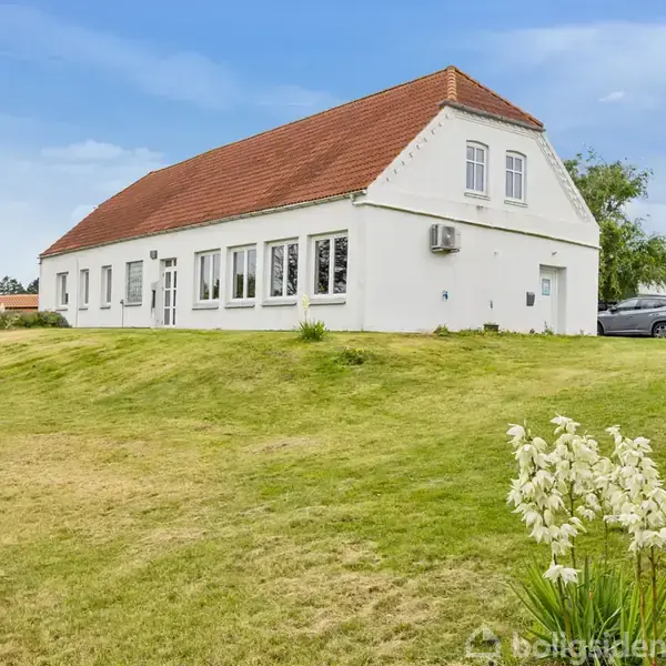 Hvidt hus med rødt tag står på en grøn græsplæne. Bil parkeret til højre. Blå himmel og blomster i forgrunden.