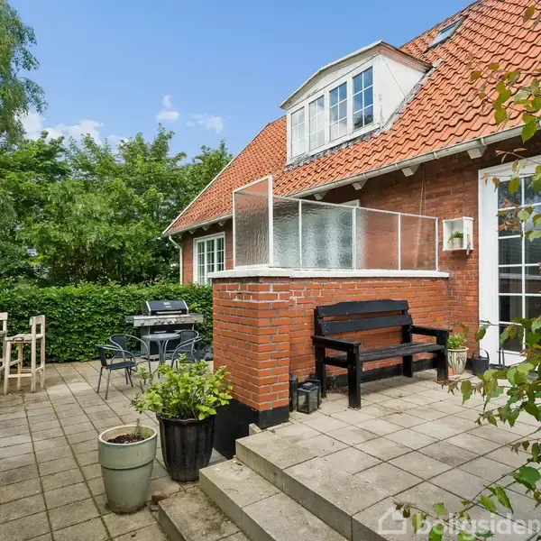 Hus med rødt tegltag ligger i grøn have. På terrassen er der sorte og træ havemøbler, krukker med planter, og en trappe fører op til en sort bænk og en glasdør.