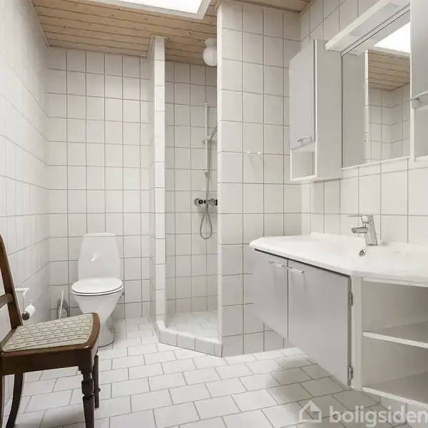 En stol står på et badeværelse. Rummet har hvide fliser, en bruser i hjørnet, et toilet ved siden af og en vask med spejl samt skabsplads til højre.