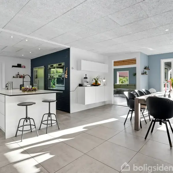 Køkken med hvid ø og to barstole, vaser og en kurv med frugt på bordet, sollys fra store vinduer, spisestue ved siden i en moderne, åben rumindretning.