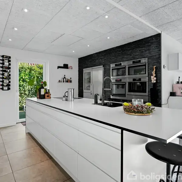 Et moderne køkken med en stor, hvid køkkenø. Barstole er placeret ved øen. Baggrunden har sort væg med indbyggede ovne og en glasdør, der fører ud til en have.