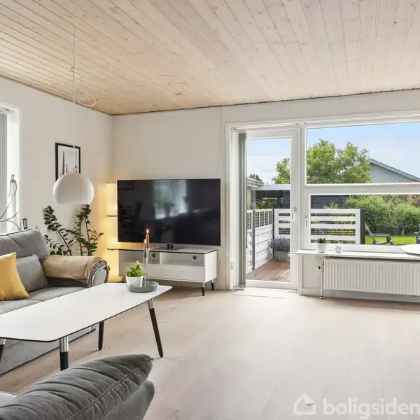 Stue med grå sofa og hvidt sofabord, en stor fladskærm på TV-bord. Vinduer afslører en terrasse og have med grønne buske. Træloft og lyse trægulve giver rummet et lyst udtryk.