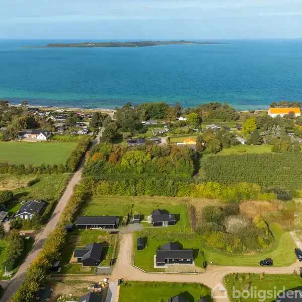 Luftfoto af et kystområde med flere huse i forgrunden og grønne marker. En vej snor sig gennem området, og i baggrunden ses en blå havoverflade med en ø i horisonten.