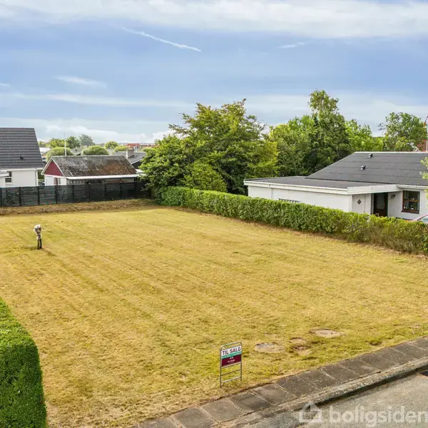 En rektangulær græsplæne med en hæk på tre sider. Plænen er omgivet af huse og træer i et beboelsesområde. Et skilt med teksten “14A, KLAGSMOSEVEJ” står foran græsplænen.