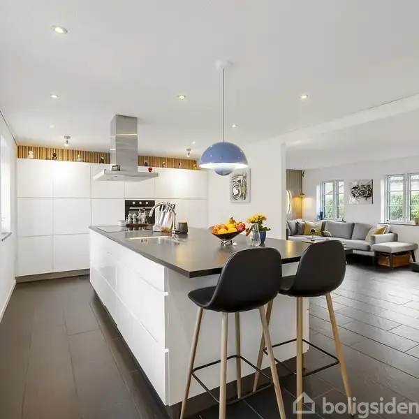 Moderne køkken med hvidt køkkenbord, to sorte barstole og frugtskål. I baggrunden ses en stue med sofa og flere vinduer, der giver naturligt lysindfald.
