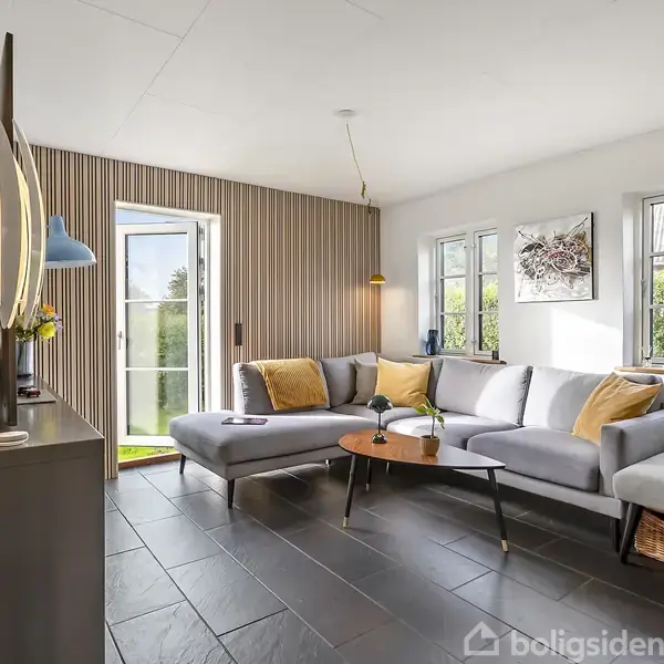 Moderne stue med en grå hjørnesofa og gule puder, placeret på mørkt flisegulv. En åben dør giver udsigt til en grøn have, mens lamper og dekorationer tilføjer hygge.