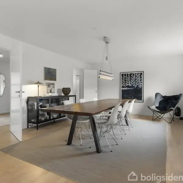 Et langt træbord med hvide stole står i en moderne spisestue. Væggene er hvide, og et maleri hænger på væggen, mens store vinduer giver naturligt lys.