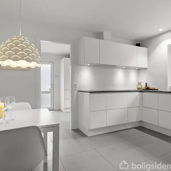 Hvidt køkken domineret af minimalistiske skabe og en sort bordplade. Et spisebord til venstre med vin og glas. Udsigt til en grøn have gennem vinduet til højre.