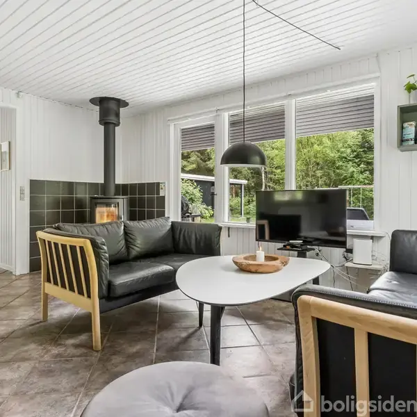 Stue med sorte lædersofaer omkring et rundt hvidt bord. Brændeovn i hjørnet, væghængt lampe over bordet. Vinduer viser grøn udsigt. Flisegulv og hvidmalede vægge.