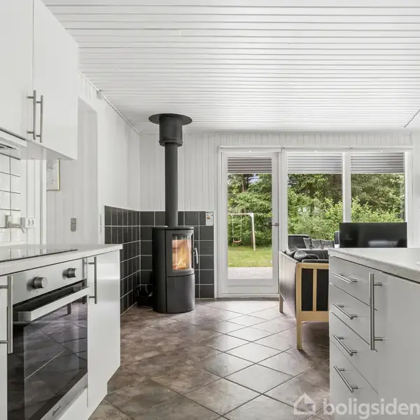 Køkken med hvidt interiør har en brændeovn ved siden af glasdøre ud til en have. Fliser på gulvet og hvide vægge skaber et lyst og modernistisk miljø.