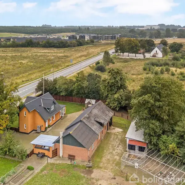 Et landhus med stråtækt tag står omgivet af flere træer og en stor græsplæne. Baggrunden viser en landevej og åbne marker under en klar himmel.