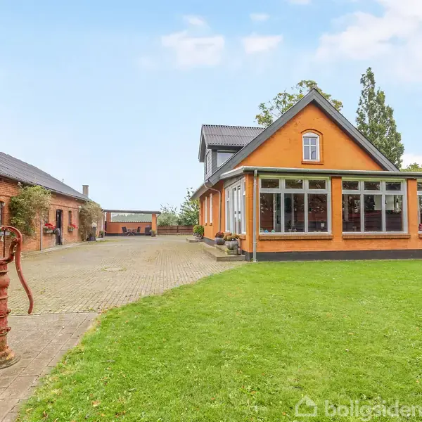Gult hus med stor veranda står på grøn plæne. Til venstre ses en gammel vandpumpe og rødt murstenshus. Skyfri himmel i baggrunden.