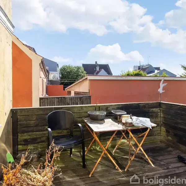 En lille altan har et bord og en stol placeret ved siden af en mur. Bordet er dækket med diverse genstande. Omkring er der bygninger og grønne træer under en blå himmel.