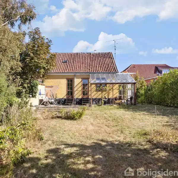 Et gult hus med rødt tegltag har en overdækket terrasse i haven. Terrassen er omgivet af grønne buske og træer under en lys blå himmel med skyer.