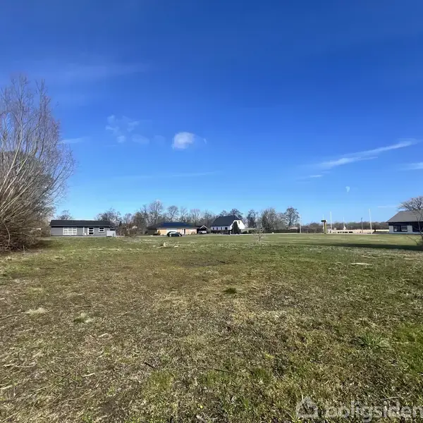 Græsareal strækker sig foran enkeltstående træer og enkelte huse i baggrunden, under en blå himmel med få skyer.