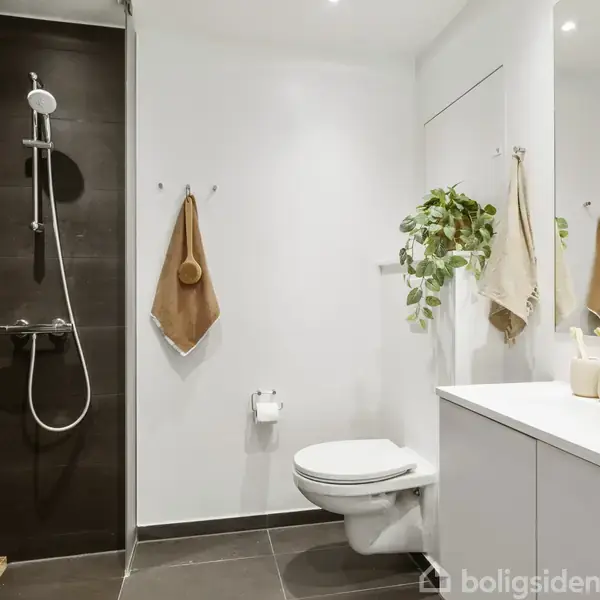 Et moderne badeværelse med en bruser til venstre, en væghængt toilet midtfor, og en hvid vask til højre. Vaskemaskine og tørretumbler er stablet i et hjørne.