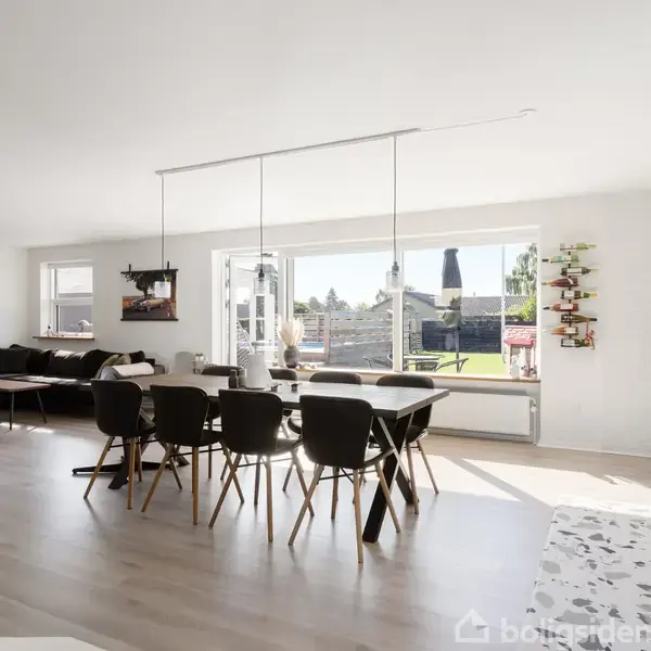 En spisestue med et langt bord og sorte stole står centralt. Store vinduer slipper lys ind, og der er adgang til en terrasse. Rummet er lyst og moderne indrettet.