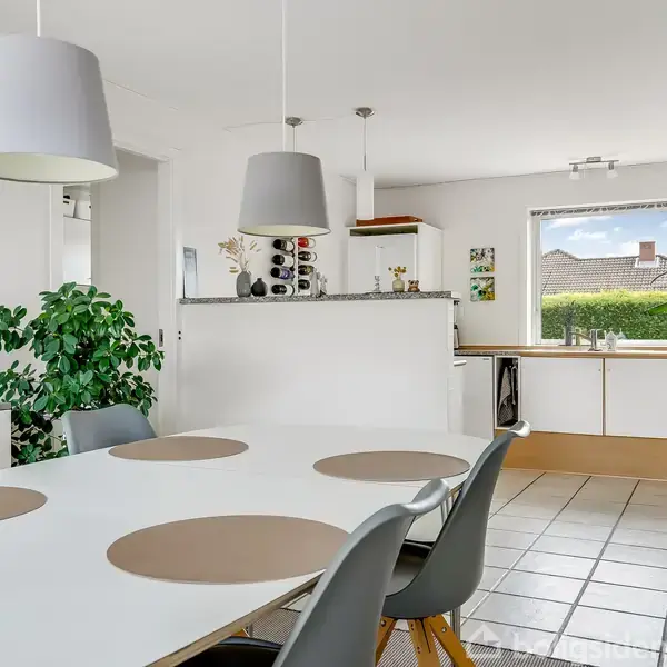 Et hvidt spisebord med grå stole og runde dækkeservietter står i et lyst køkkenalrum. Grønne planter og moderne lamper dekorerer rummet. En vindue viser villaudsigt.