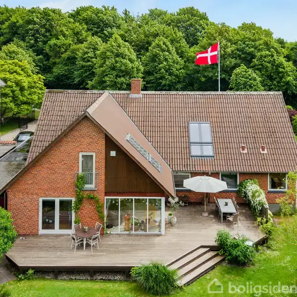 Et rødt murstenshus med et flag vajende fra taget. Foran huset er en rummelig træterrasse med havemøbler og parasol. Grønt område og skov danner baggrunden.
