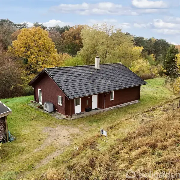 Et rødt træhus med sort tag står i en frodig, grøn landskab med efterårsfarvede træer omkring. En lille bygning er ved siden af grusstien, der leder til huset.
