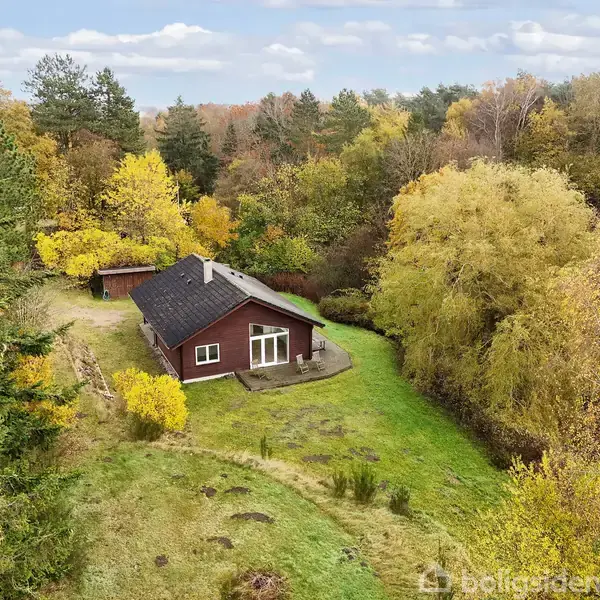 Et rødt hus med mørkt tag står midt i en lysning omgivet af tæt efterårsskov med farverige træer under en skyet himmel.