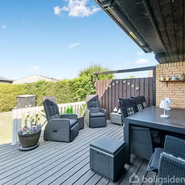 Terrasse med sorte kurvemøbler arrangere omkring et rektangulært bord, omgivet af en velplejet have med hække. En murstensvæg med hylder præger baggrunden, og himlen er klar med få skyer.