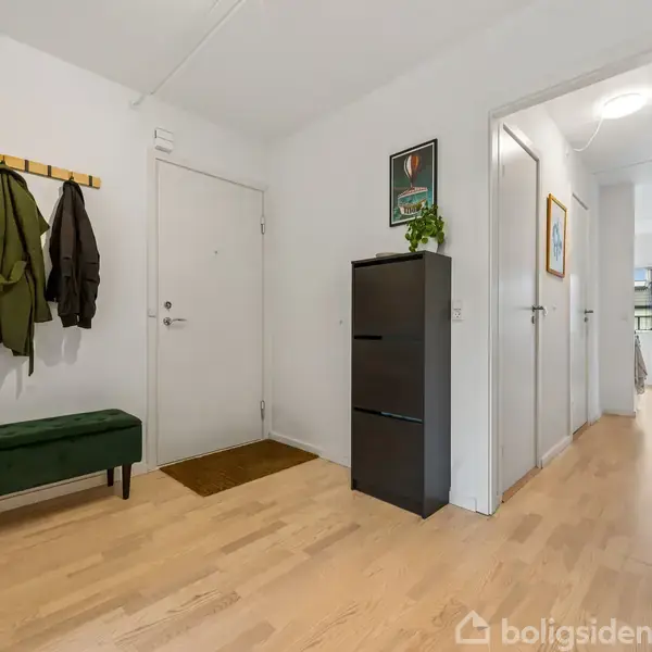 Gang med lyst trægulv, en grøn bænk og knagerække med jakker. En sort skænk med plante står ved siden af en lukket dør; en åben gang fører til andre rum.
