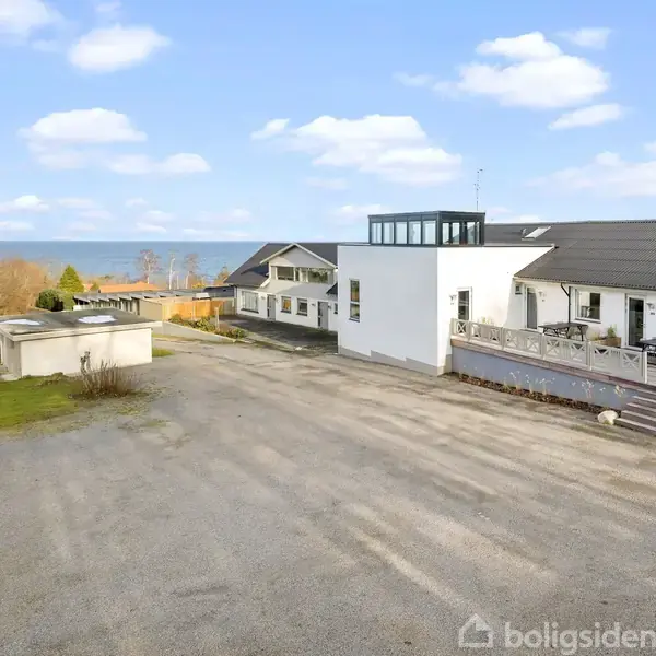 En række huse med fladt tag og store vinduespartier står stille langs en bred, grusbelagt indkørsel. I baggrunden ses havet og en skovklædt kystlinje.