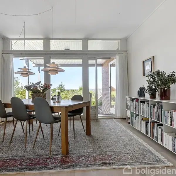 Et spisebord med stole står på et tæppe i en lys spisestue. Store vinduer viser udsigt til natur udenfor. En reol med bøger og en lampe står ved siden.