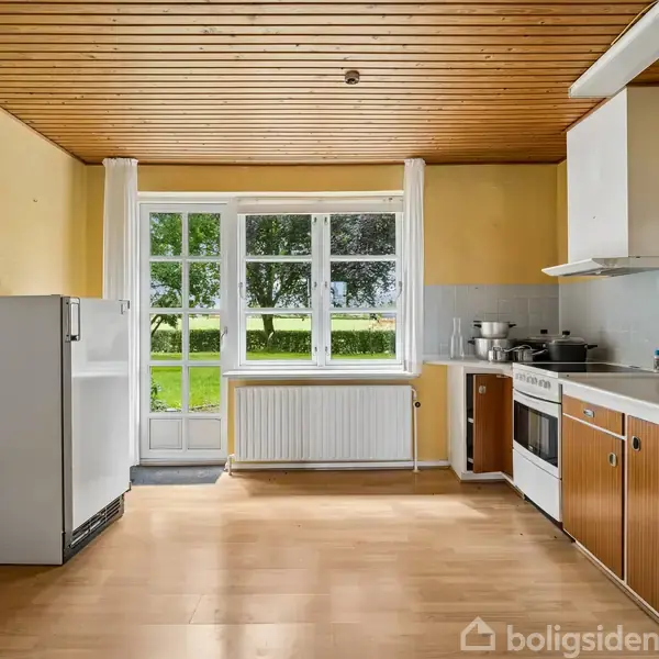 Køkken med træloft og gule vægge. Hvidt køleskab og komfur med emhætte står ved trækøkkenborde. Store vinduer giver udsigt til grøn have. Radiator under vindue. Ingen tekst i billedet.