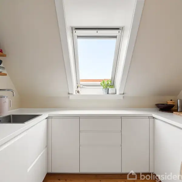 Hvidt, moderne køkken med vinkelformet bordplade. Et ovenlysvindue slipper naturligt lys ind, og en plante står nederst. Redskaber hænger på væggen ved siden af en indbygget ovn.