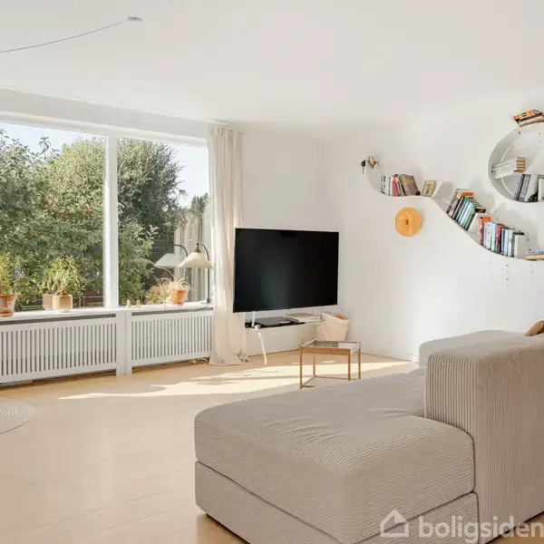 Stue med grå sofa, rund væghylde med bøger, og stor fladskærms-TV. Stort vindue med udsigt til have og flere planter på vindueskarmen. Lyst trægulv og gyngestol i hjørnet.