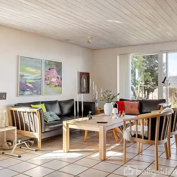 Stue med sorte og træfarvede møbler, centralt placeret sofabord fyldt med lysestager. Kunst tilføjer farve på væggen. Store vinduer bader rummet i dagslys.