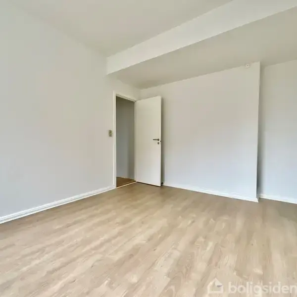 Et tomt rum med lyst trægulv, en hvid dør, og hvide vægge, som skaber en minimalistisk atmosfære. Døren står åben og fører til en anden del af huset.