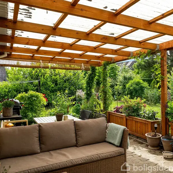 Sofa står under en træpergola med gennemsigtig tagdække. Haven i baggrunden er frodig med buske og blomster, mens potter og et havebord ses omkring terrassen.