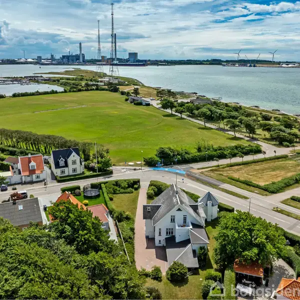 Hus omgivet af grønne haver ved kysten, med udsigt over vandet og fladt græsareal. I baggrunden ses industriområder og vindmøller under en blå himmel.
