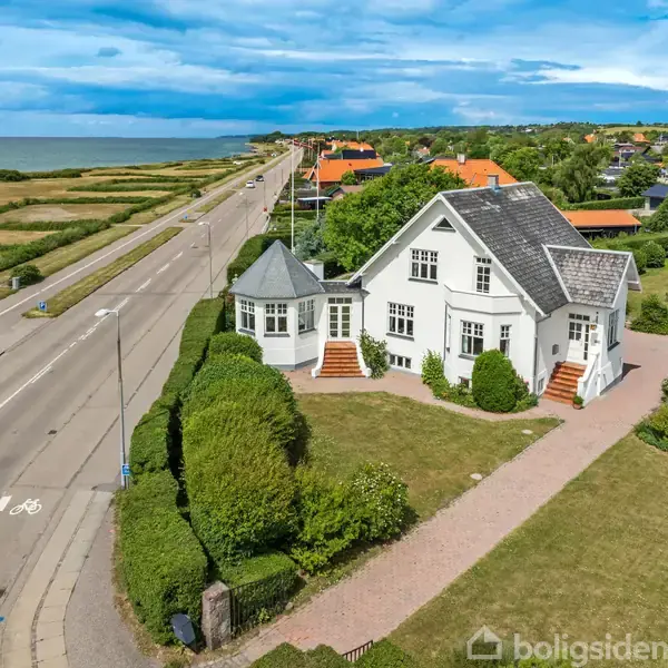 Hvid villa med sort tag står på hjørnegrund ved en kystvej. Den har trapper, hæk omkranser haven. Baggrunden viser havet til venstre og flere huse til højre.
