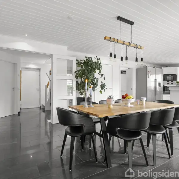Et stort spisebord af træ med seks sorte stole står i et moderne spisestuemiljø. Rummet har hvide vægge, sorte gulvfliser, og en plante i hjørnet samt hængelamper over bordet.