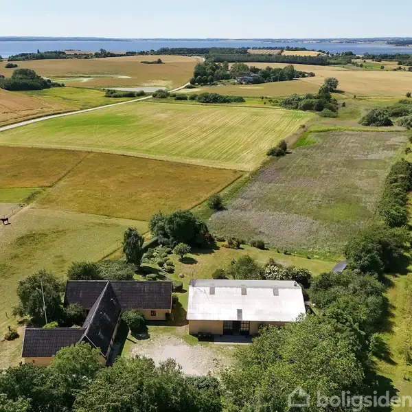 Gårdbygninger ligger midt i åbne marker med spredt bevoksning. Omkringliggende landskab består af varierede græsarealer og små skovområder, med udsigt over en fjern kystlinje i horisonten.
