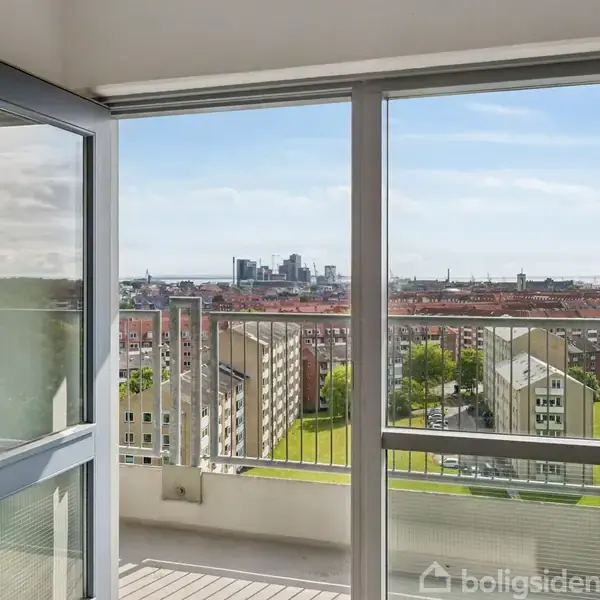 Åben altandør ud mod solbeskinnet byudsigt med moderne bygninger og grønne områder i baggrunden. Høje boliger ses under en klar blå himmel.