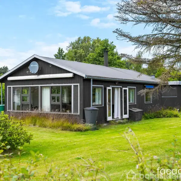 Sort træhus med store vinduer står fredeligt i en grøn have omkranset af træer og buske. Blå himmel med få skyer ses i baggrunden.