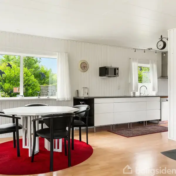 En rund hvid bordgruppe med sorte stole står på et rødt tæppe i en lys, moderne stue. Et brændeovn varmer rummet, der er omgivet af vinduer med udsigt til grønne planter.