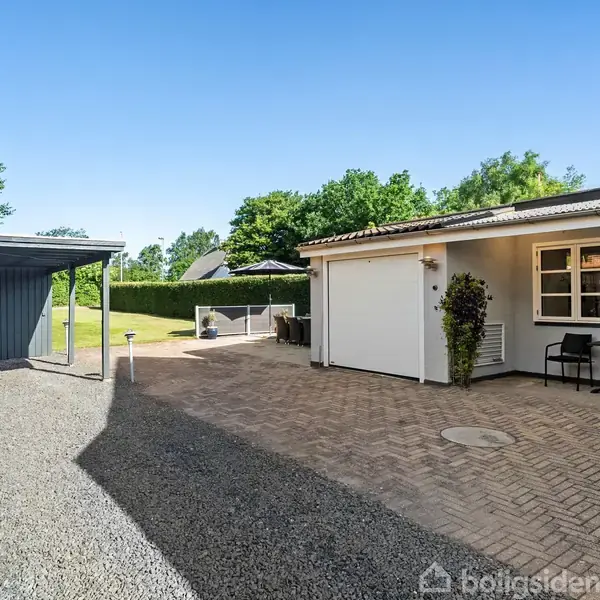 Et hvidt hus med en garage til højre og en indkørsel med grus og fliser. Til venstre er en sort carport, omgivet af grønt græs og træer under en klar blå himmel.