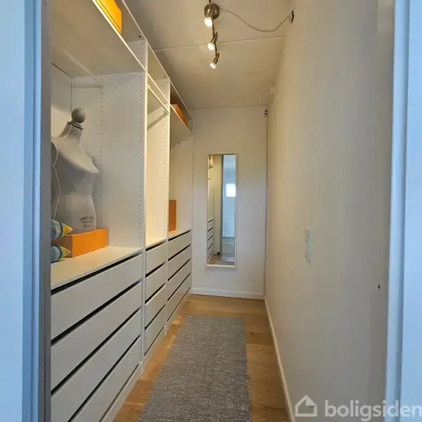 Mannequin står blandt hvide skabe med skuffer i en garderobe. En gulvkrog ligger på trægulvet. Et spejl hænger på endevæggen. Lys strømmer ind fra vinduet.