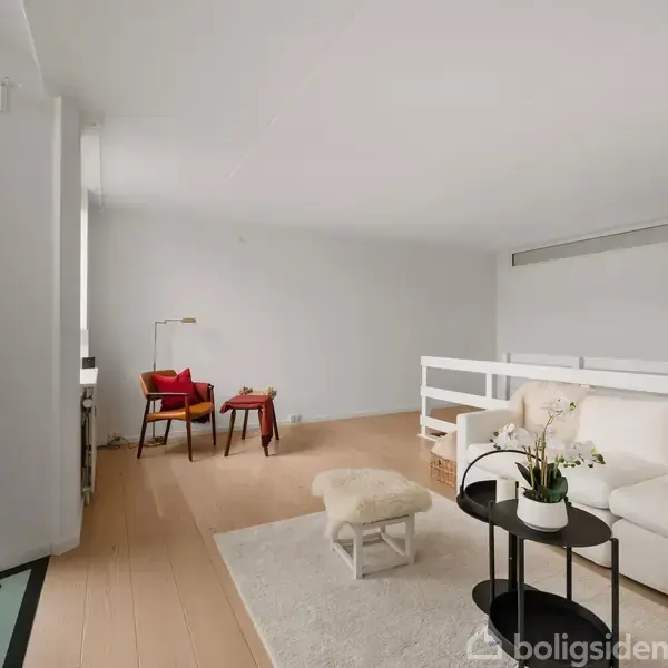 En moderne stue med en hvid sofa og et sofabord, placeret på et lyst trægulv. I baggrunden står to røde stole ved et skrivebord med en lampe.